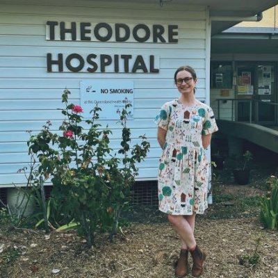 A woman in a dress and boots stands in front of a building which is labelled Theodore Hospital